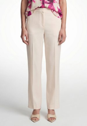 Woman wearing beige straight-leg trousers, open-toe beige heels, and a floral pink blouse, standing against a white background.