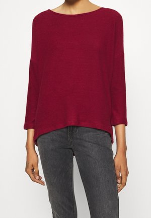 Person trägt einen locker sitzenden, langärmligen burgunderfarbenen Pullover mit Bootsausschnitt und dunkelgraue Jeans und steht vor einem weißen Hintergrund.