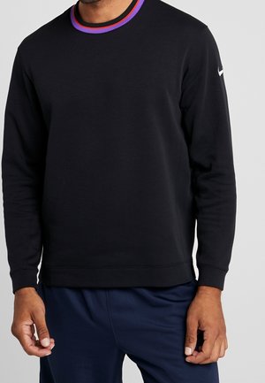 Homme portant une chemise noire à manches longues avec un col rayé violet et rouge et un pantalon bleu marine, debout avec les mains détendues.
