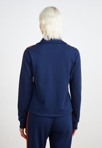 Marinblå träningsoveralljacka med krage, med orange sidestreck och långa ärmar, tillverkad av ett slätt tyg.