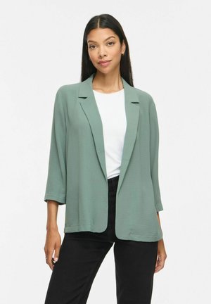 Femme aux longs cheveux foncés portant un blazer vert clair ouvert sur une chemise blanche et un pantalon noir, debout devant un fond blanc uni.