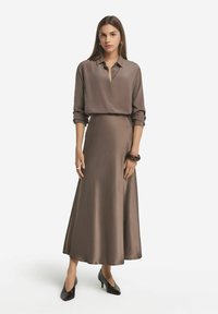 Braunes Satin-Kleid mit einem kragenlosen Hemdoberteil und einem fließenden Rock. Glatte Textur, lange Ärmel hochgekrempelt, kombiniert mit schwarzen spitzen Schuhen.