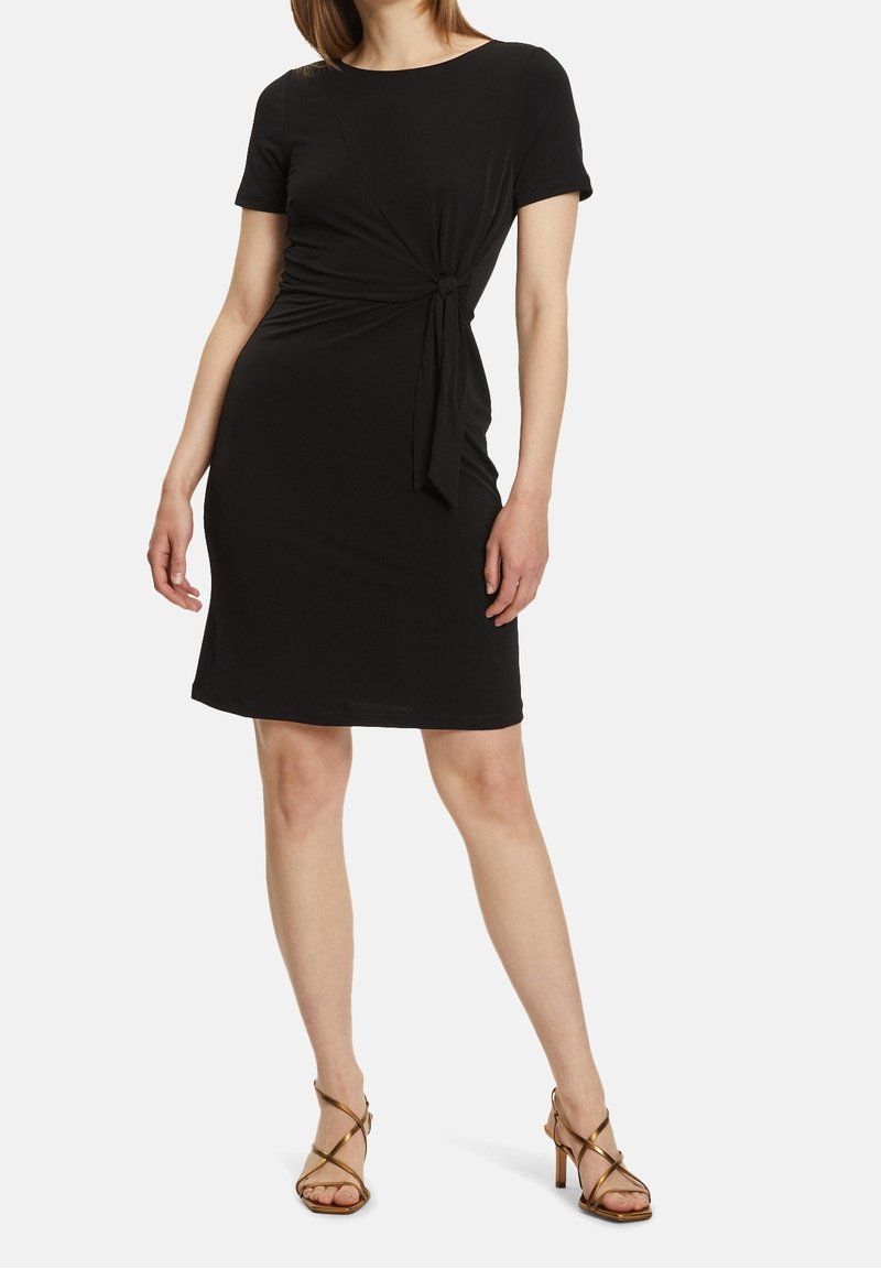 Femme portant une robe noire mi-longue à manches courtes avec un nœud sur le côté, associée à des sandales à talons hauts bronze à brides.