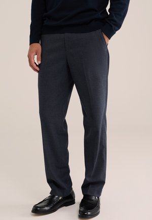 Homme portant un pantalon droit gris foncé avec des mocassins en cuir noirs, une main dans la poche, debout sur un fond beige uni.