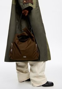 Brązowa torba z materiału o zaokrąglonym kształcie, z dwoma bocznymi suwakami i uchwytem u góry. Zawiera logo oraz teksturowane wykończenie. Nosi ją w połączeniu z szerokimi beżowymi spodniami.