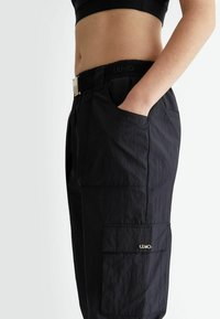 Pantaloni cargo neri realizzati in tessuto leggero, con vita cinturata, tasche laterali e un accento logo dorato sulla tasca cargo.