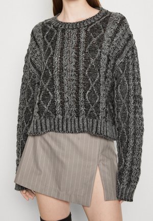 Mujer con un jersey gris grueso de punto trenzado y una minifalda beige de rayas con abertura delantera, mostrada desde los hombros hasta mitad del muslo.