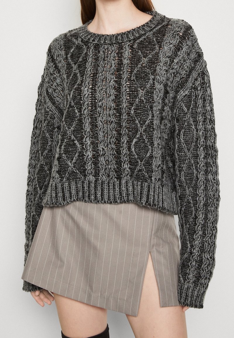 Femme portant un pull épais gris torsadé et une mini-jupe beige à rayures avec une fente devant, vue des épaules jusqu'au milieu de la cuisse.