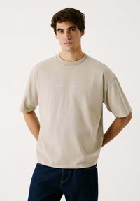 Beige Baumwoll-T-Shirt mit Rundhalsausschnitt und kurzen Ärmeln, das einen dezenten "RED BULL RACING" Druck in hellem Text über der Brust trägt.