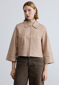 Trumpas šviesiai rožinės odos švarkas su priekyje užtrauktuku, apykakle ir klostuotu apačios kraštu. Turi platėjančias rankoves ir šonines kišenes, dėvimas ant tamsių kelnių.