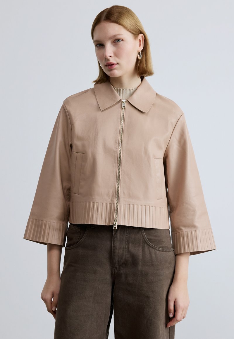 Trumpas šviesiai rožinės odos švarkas su priekyje užtrauktuku, apykakle ir klostuotu apačios kraštu. Turi platėjančias rankoves ir šonines kišenes, dėvimas ant tamsių kelnių.