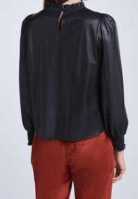 Svart läderblus med hög volangkrage, nyckelhålsbak och puffärmar; har texturerad svart smockning vid ärmsluten, ihop med röda kordbyxor.