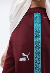 Μπορντό αθλητικό σορτς με κάθετη τυπωμένη τιρκουάζ ρίγα, λευκό λογότυπο Puma και κείμενο "KING", φορεμένα από άτομο που σηκώνει λευκό πουκάμισο.