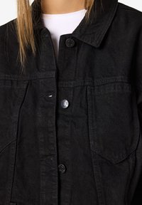 Black denim jacket with metal buttons, chest pockets, and visible stitching. The fabric has a textured finish, emphasizing durability and style.