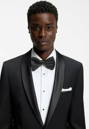 Smoking nero con rever in raso, camicia bianca plissettata e papillon nero testurizzato. Fazzoletto da taschino bianco, bottoni neri. Semplice ed elegante.