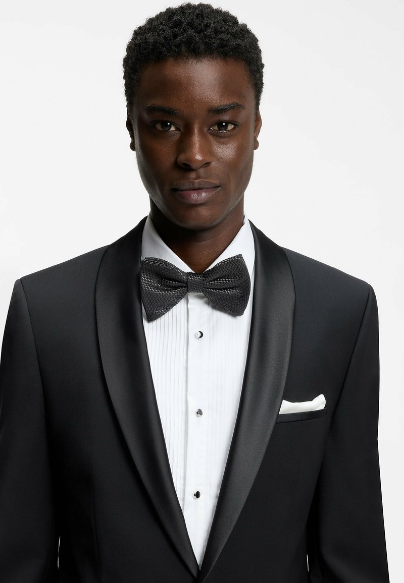 Czarny smoking z satynowymi klapami, biała plisowana koszula i teksturowana czarna mucha. Poszetka jest biała, guziki czarne. Prosty i elegancki.