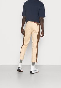 Beiga byxor med bruna sidopaneler, kort längd och bakfickor. Matchas med vita högklackade sneakers och grå strumpor, bärs med en mörk skjorta.