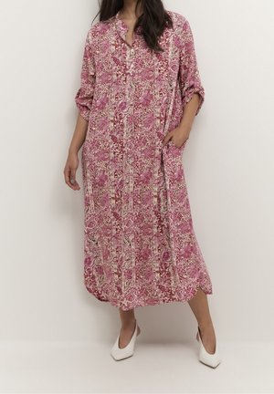 Frau trägt ein lockeres, langes rosa-weißes Kleid mit Blumenmuster, hochgekrempelten Ärmeln und weißen spitzen Absätzen und steht vor einer schlichten Wand.