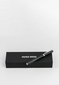 Črn kovinski pisalo s kromiranim sponko in dodatki, postavljeno poleg pravokotne črne škatle, na kateri je v beli barvi natisnjeno "HUGO BOSS".