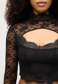 Svart långärmad topp med blommigt spetsmönster, hög krage, utskuret detaljer och vågad nederkant. Halvgenomskinlig textur överallt.