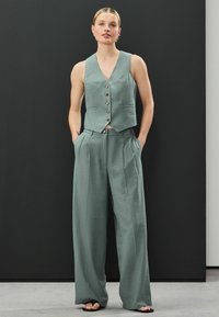 Hellgrünes Leinenweste mit dekorativen Knöpfen, kombiniert mit passenden weit geschnittenen Hosen. Taschen in den Hosen, minimale Textur überall.
