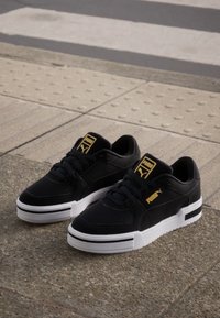 Black sneakers with a smooth leather upper, white rubber sole, gold logo accents, and a low-top design. Features perforated details on the toe.