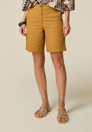 Donna che indossa pantaloncini giallo senape fino al ginocchio e sandali intrecciati con lacci alla caviglia, in piedi davanti a uno sfondo semplice.