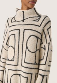 Beige tröja med hög hals, med svarta geometriska mönster. Mjuk textur, något överdimensionerad passform och distinkta vinklade design detaljer.