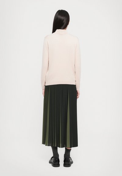 Woman with long dark hair wearing a light pink sweater and a black and olive-green pleated midi skirt, standing facing away on a plain background.