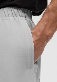 Pantalones cortos gris claro hechos de tela texturizada, con una cinturilla relajada y un bolsillo lateral visible cerca de una mano. Patrón de cuadrícula sutil.