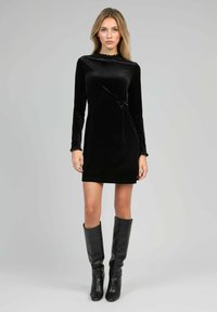 Femme portant une robe mini à manches longues en velours noir avec un nœud sur le côté, se tenant devant un fond gris uni, assortie de bottes noires jusqu'aux genoux.