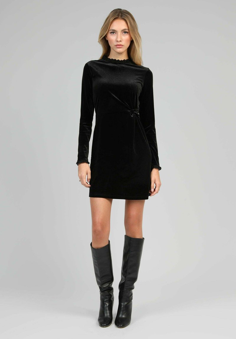 Femme portant une robe mini à manches longues en velours noir avec un nœud sur le côté, se tenant devant un fond gris uni, assortie de bottes noires jusqu'aux genoux.