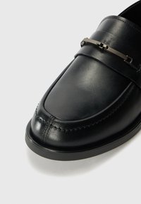 Pantofi loafer din piele neagră, cu talpă plată, cusături evidente și un accent metalic pe partea superioară. Textură netedă și design elegant.