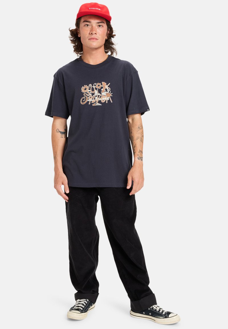 Jauns vīrietis valkā sarkanu cepuri, tumši zilu grafisku T-kreklu, melnas bikses un melnus sporta apavus, stāvēdams ar relaksētu poza un skatoties uz priekšu.