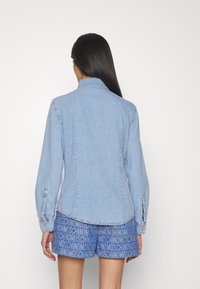 Ljusblå denimjacka med långa ärmar och knäppte manschetter, med en klassisk krage och figurnära silhuett. Bärs med mönstrade shorts.