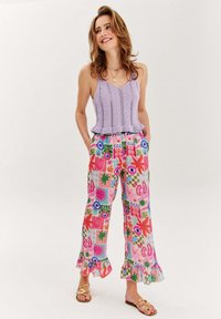 Kobieta uśmiechnięta, ubrana w lawendowy dzianinowy top na ramiączkach, kolorowe szerokie spodnie w wzory z falbanami i złote sandały na paski, stoi z rękami w kieszeniach.