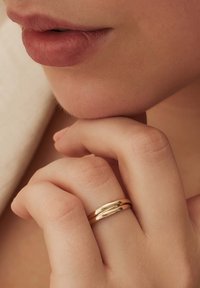 Goldene Doppelring auf einem Finger, mit einer glatten, polierten Oberfläche und sanfter Kurve, vor einem weichen neutralen Hintergrund.