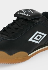Μαύρο αθλητικό παπούτσι Umbro με λευκό λογότυπο διπλού διαμαντιού, δέσιμο με κορδόνια μπροστά και καφέ υφασμένη σόλα με σήμανση Umbro.