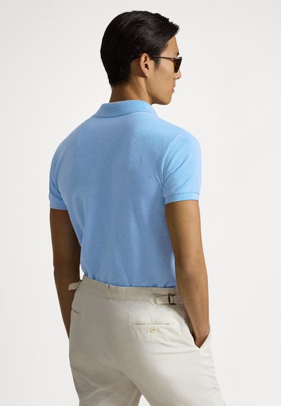 Ανοιχτό γαλάζιο polo με κοντά μανίκια και υφή πλεξίματος, με κλασικό γιακά. Συνδυασμένο με μπεζ παντελόνι, σε προβολή από πίσω.