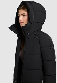 Giubbotto imbottito nero con un grande cappuccio imbottito, chiusura a zip frontale e bottoni a pressione; presenta una toppa con logo sulla manica sinistra.