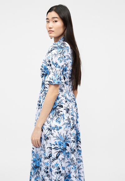 Γυναίκα με μακριά σκουρόχρωμα μαλλιά, ντυμένη με άσπρο φόρεμα με μπλε και μαύρα λουλουδάτα σχέδια, στέκεται πλάγια σε απλό φόντο.