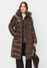 Brun puffjakke med hætte, der har horisontale quiltede sektioner, lynlås og to sidelommer. Accentueret med en suede håndtaske.