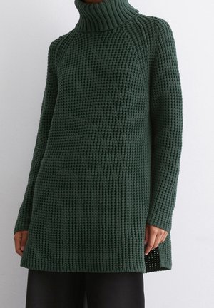 Tmavě zelený tlustý rolák se vzorem vaflového pletení a dlouhými rukávy, se spodními rozparky na bocích, nošený s černými kalhotami na jednobarevném světlem pozadí.