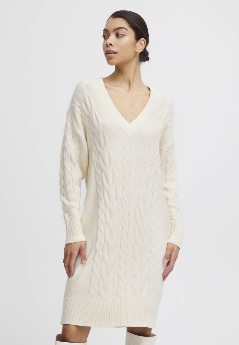 Strickkleid in Creme mit tiefem V-Ausschnitt und Zopfmuster an den Ärmeln und am Körper, abgerundet mit gerippten Bündchen und Saum.