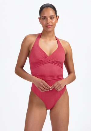 Vrouw draagt een getextureerd dieproze halterbadpak, staat met handen losjes voor zich geklemd tegen een effen witte achtergrond.