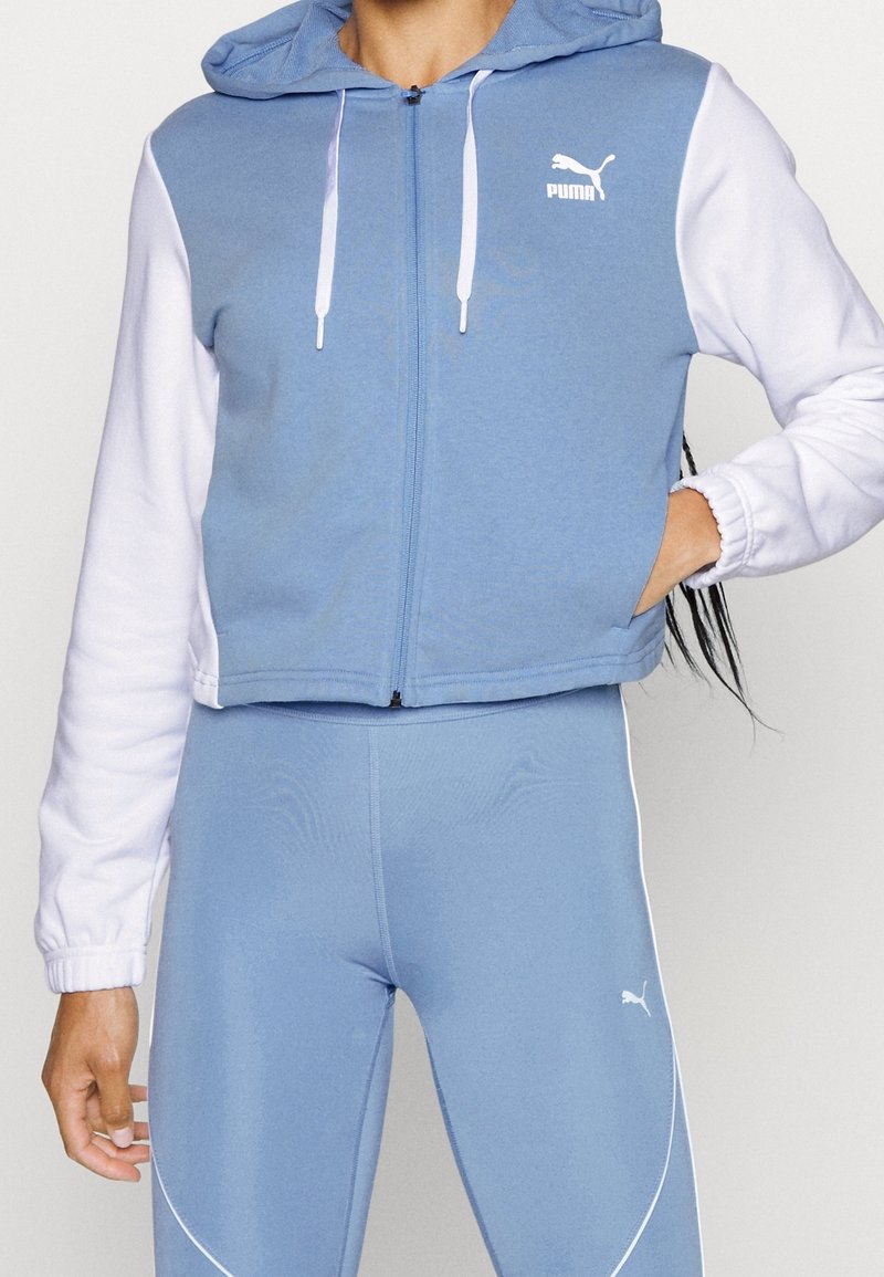 Un hoodie cropped zippé bleu clair avec des manches blanches, une capuche et le logo PUMA. Il possède une texture lisse et des poignets côtes. Accompagné de leggings assortis.