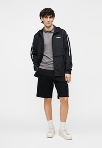 Chaqueta ligera negra con capucha, camiseta tipo polo gris, pantalones cortos negros y zapatillas beige. Presenta rayas blancas de contraste en las mangas.