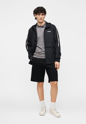 Chaqueta ligera negra con capucha, camiseta tipo polo gris, pantalones cortos negros y zapatillas beige. Presenta rayas blancas de contraste en las mangas.