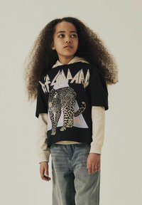 Svart grafisk t-shirt med ett leopardmönster, bärs över en ljus hoodie. Matchas med ljusblå jeans i en avslappnad passform.