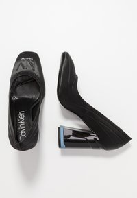 Černé vysoké podpatky s hladkým syntetickým svrškem, zaoblenou špičkou a výrazným šikmým blokovým podpatkem s modrými akcenty. Viditelné logo značky.